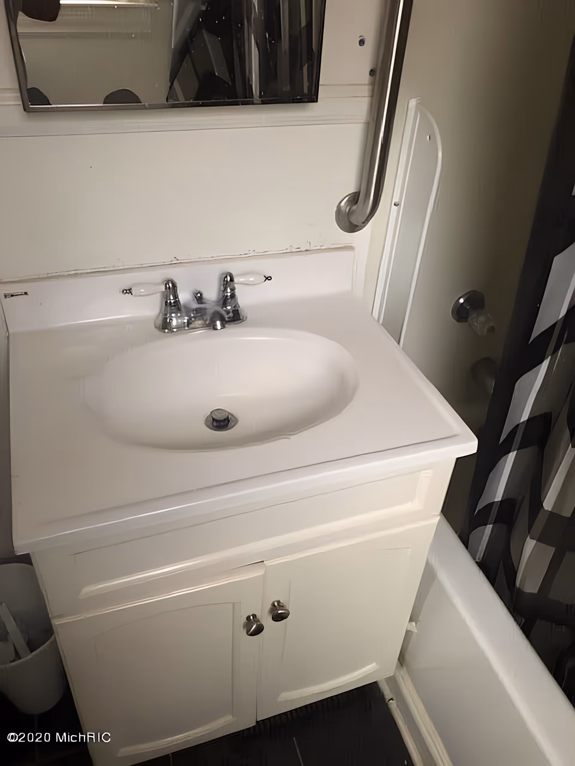 Small white bathroom vanity with sink, mirror, grab bar, and shower curtain over a bathtub.