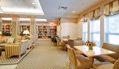 A bright and spacious common area with large windows letting in natural light. The room features several tables with chairs, each decorated with a small vase of sunflowers. There are comfortable armchairs and sofas arranged near bookshelves filled with books. The carpet has a subtle patterned design, and the walls are painted in warm neutral tones.