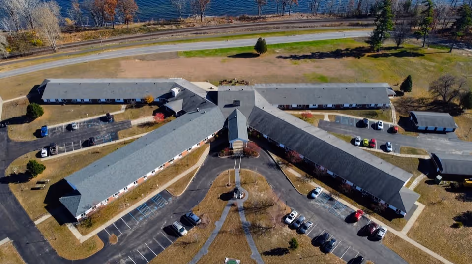Aerial view of a senior living facility with three long, connected buildings forming a Y shape. The buildings have gray roofs and red brick walls. Several cars are parked in parking lots around the buildings. The facility is situated near a body of water with a road running parallel to it. Trees with autumn foliage are visible around the property.