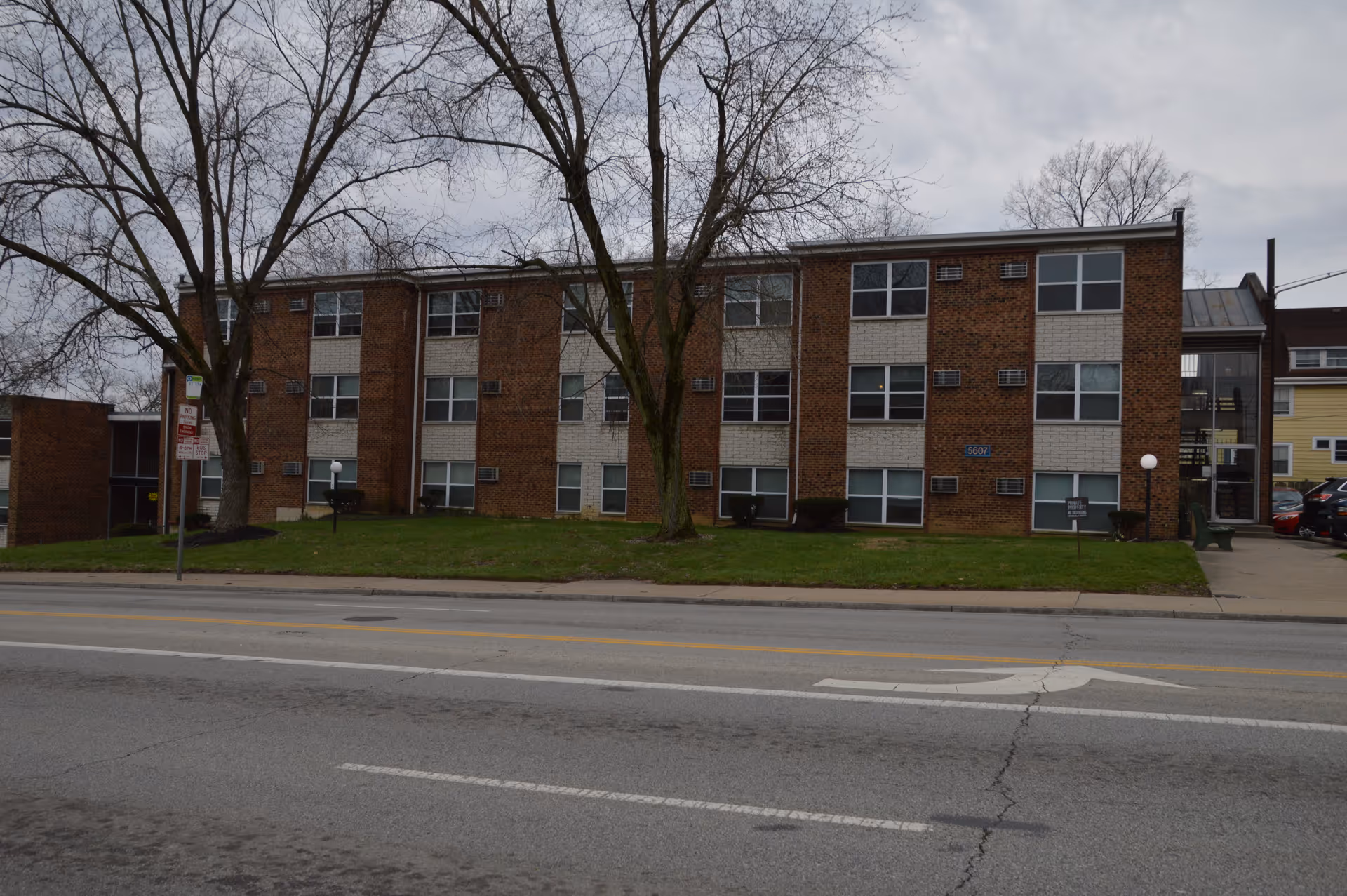Three-story brick apartment building with bare trees and a grassy lawn facing a street on an overcast day.