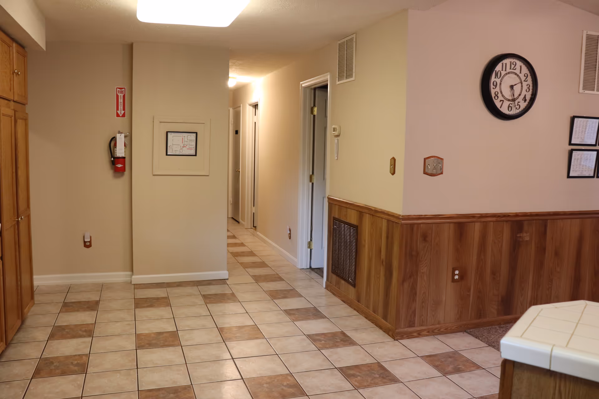 Tile-floored interior hallway with wood-paneled lower walls, a wall clock, and a fire extinguisher in an assisted living facility.