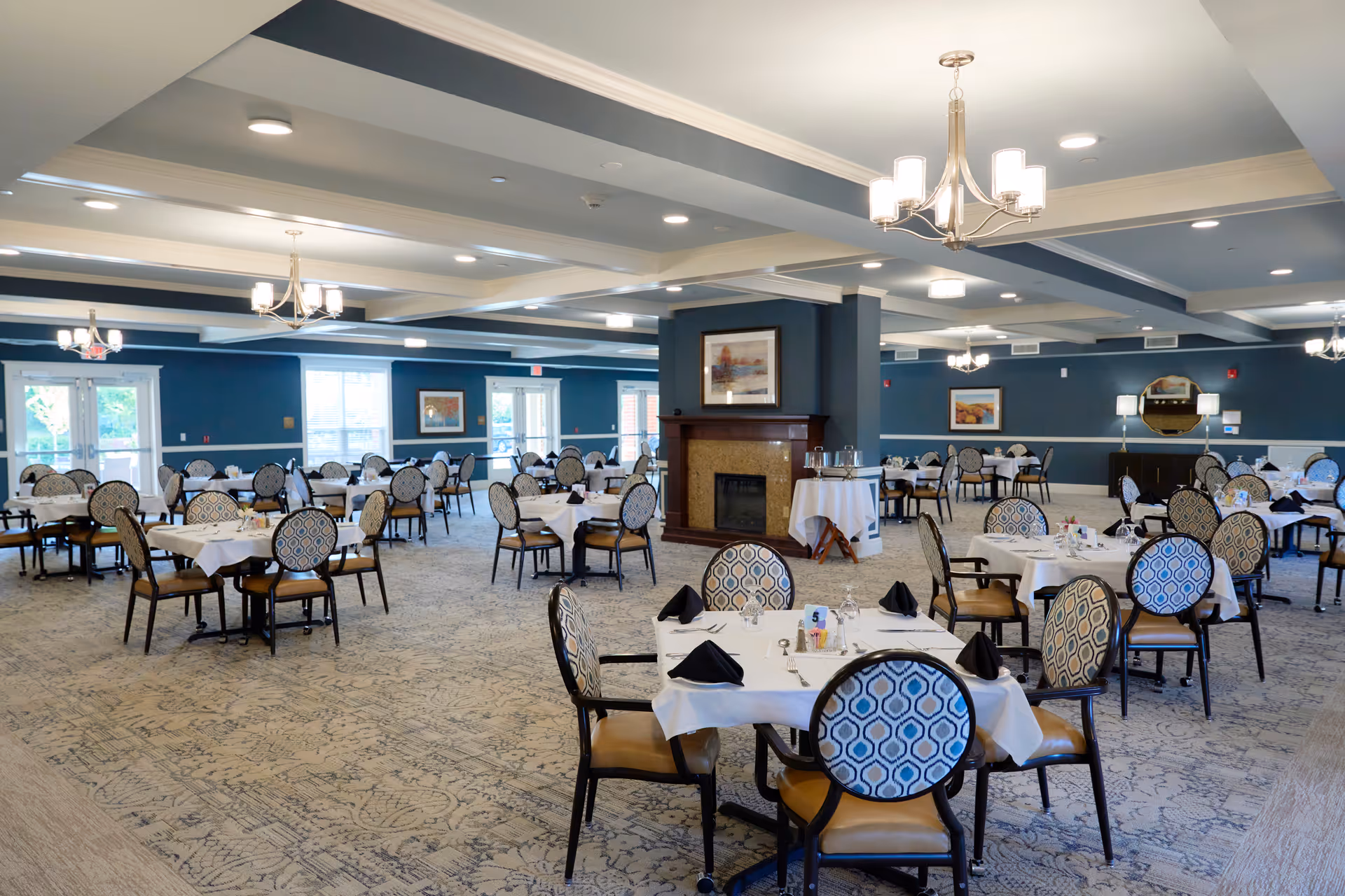 A spacious dining room with multiple round tables covered with white tablecloths, each set with black napkins, glassware, and silverware. The chairs have patterned backs and tan seats. The room features blue walls, a patterned carpet, chandeliers, and a central fireplace with a framed picture above it. Large windows and glass doors allow natural light to enter.