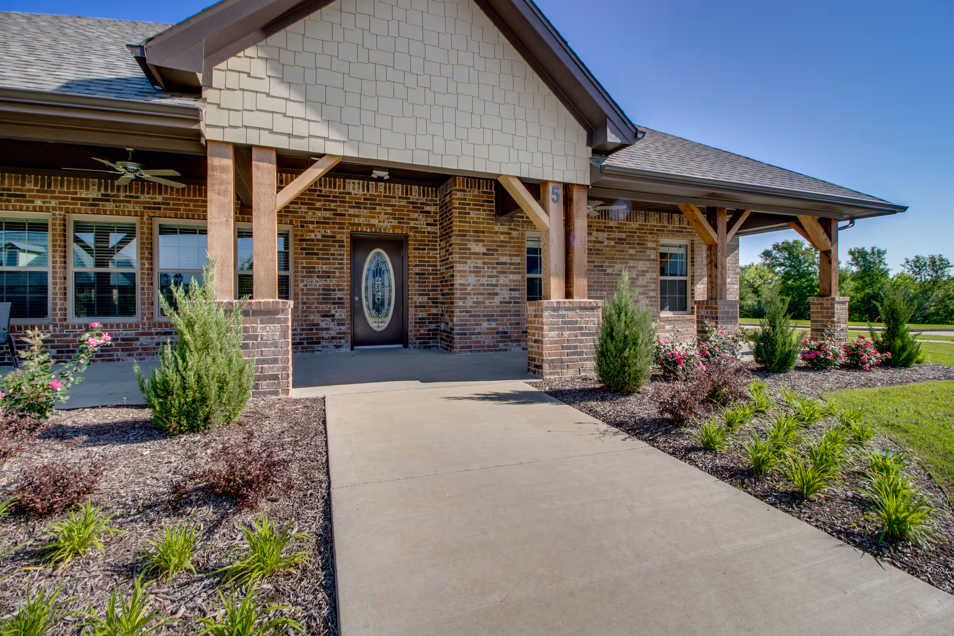 Front entrance of a brick building with a covered porch supported by wooden beams. There is a concrete walkway leading to a decorative oval glass door. The landscaping includes green shrubs and flowering plants on either side of the walkway under a clear blue sky.