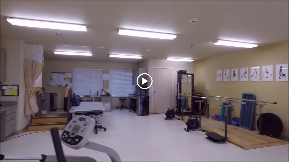 Interior view of a rehabilitation therapy room with exercise equipment including parallel bars, weights, and exercise machines. There are instructional posters on the wall demonstrating exercises, a treatment table, desks, and chairs near windows with blinds.