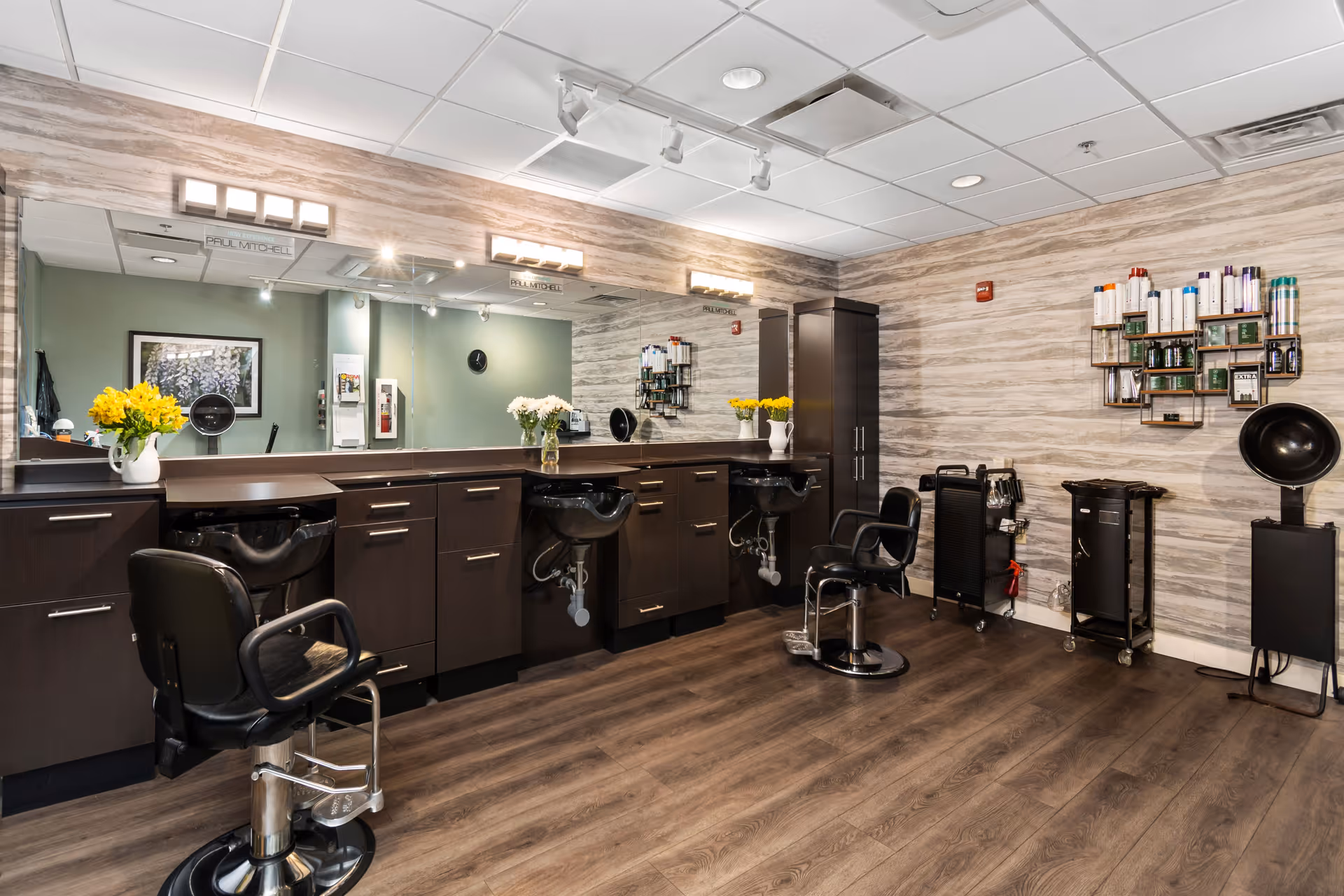 Interior of a modern hair salon with two black salon chairs in front of a long counter with sinks and large mirrors. The walls have a light wood panel design, and shelves with hair care products are mounted on the wall. The floor is dark wood, and there are vases with yellow and white flowers on the counter.