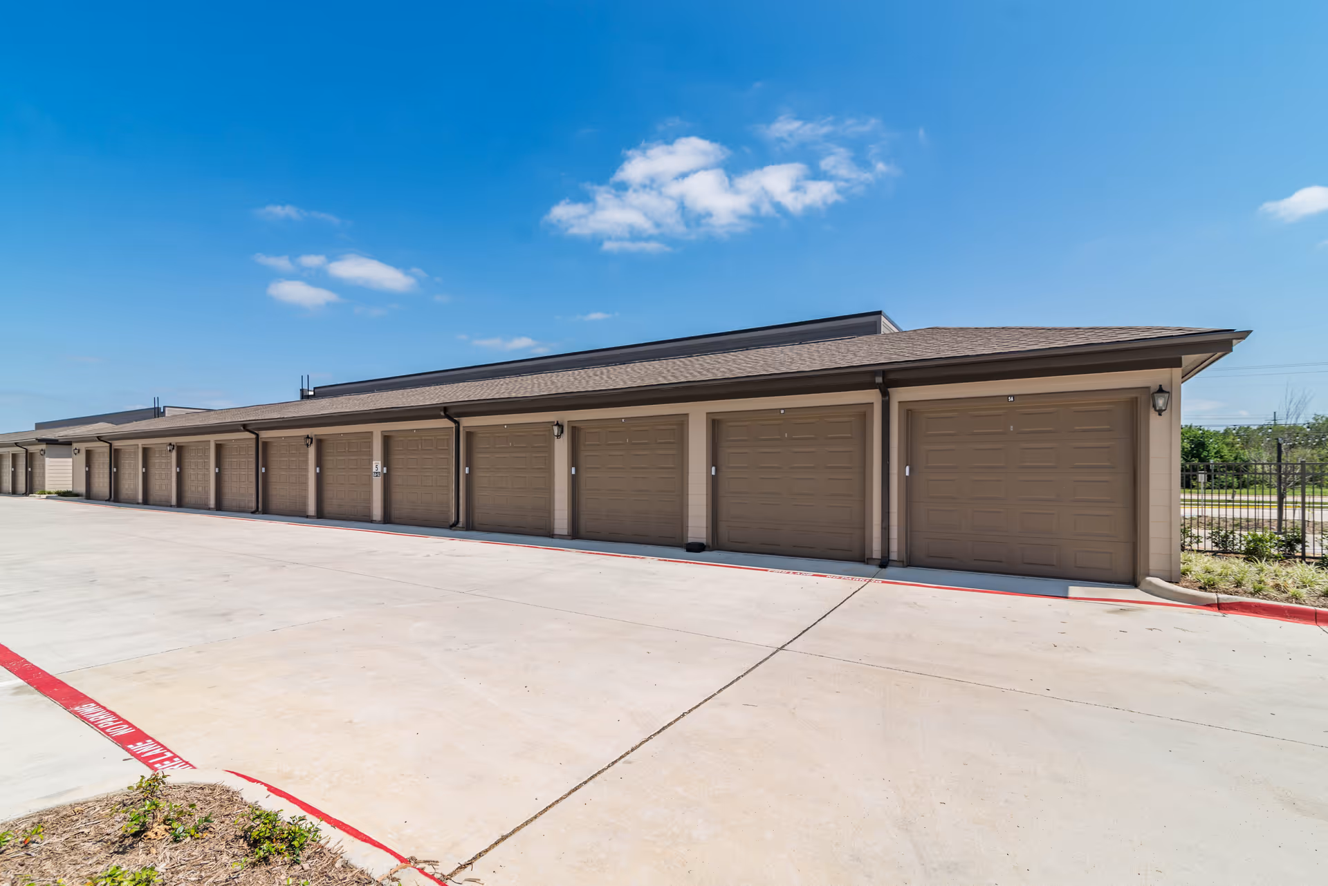 A row of closed brown garage doors attached to a single-story building under a clear blue sky with a few clouds. The garages are lined up along a wide concrete driveway with a red curb marked 'FIRE LANE NO PARKING'.