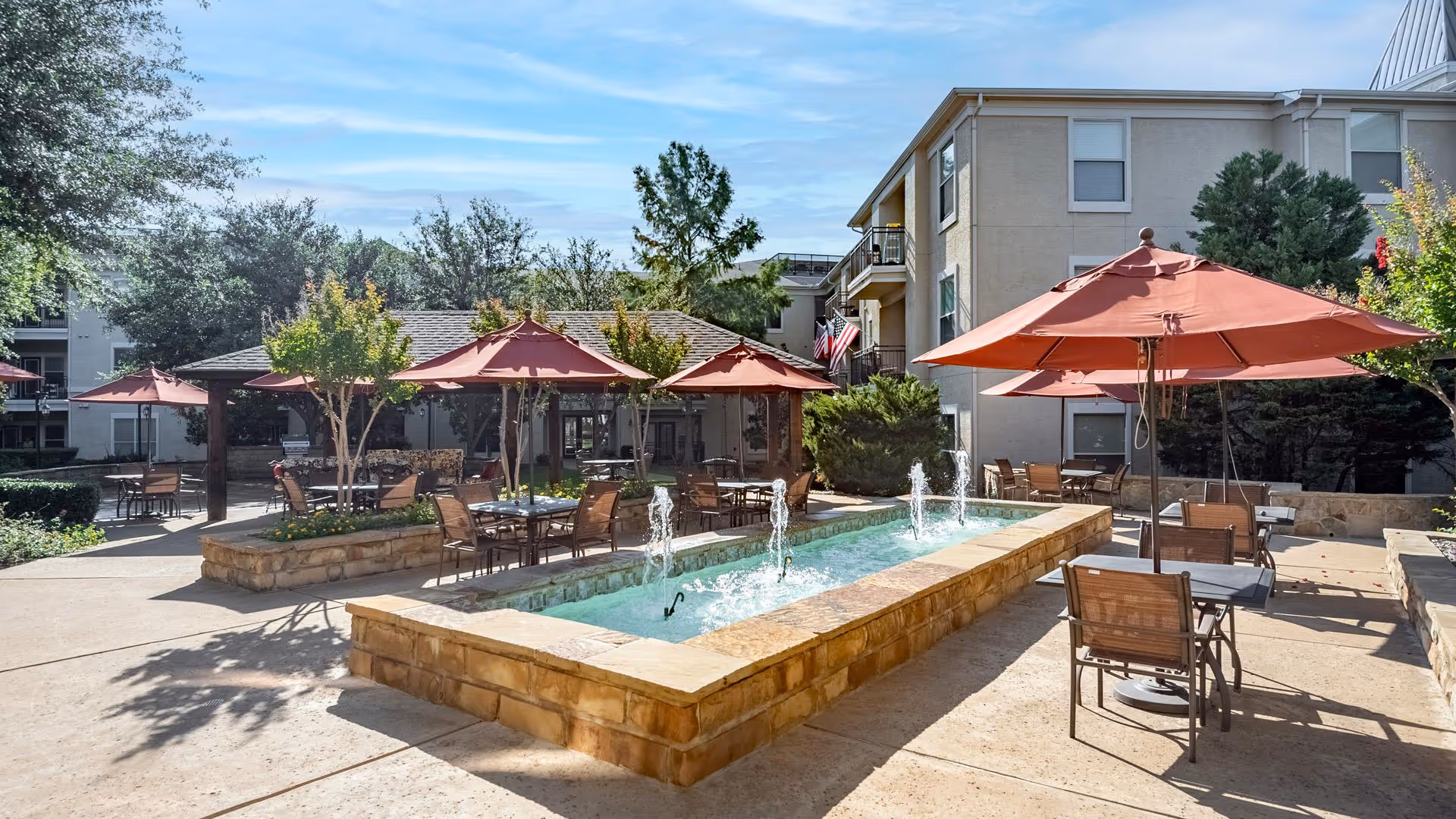 Outdoor patio area at a senior living facility with multiple tables and chairs under large red umbrellas surrounding a rectangular water fountain with several small water jets, trees, and a multi-story building in the background under a blue sky.