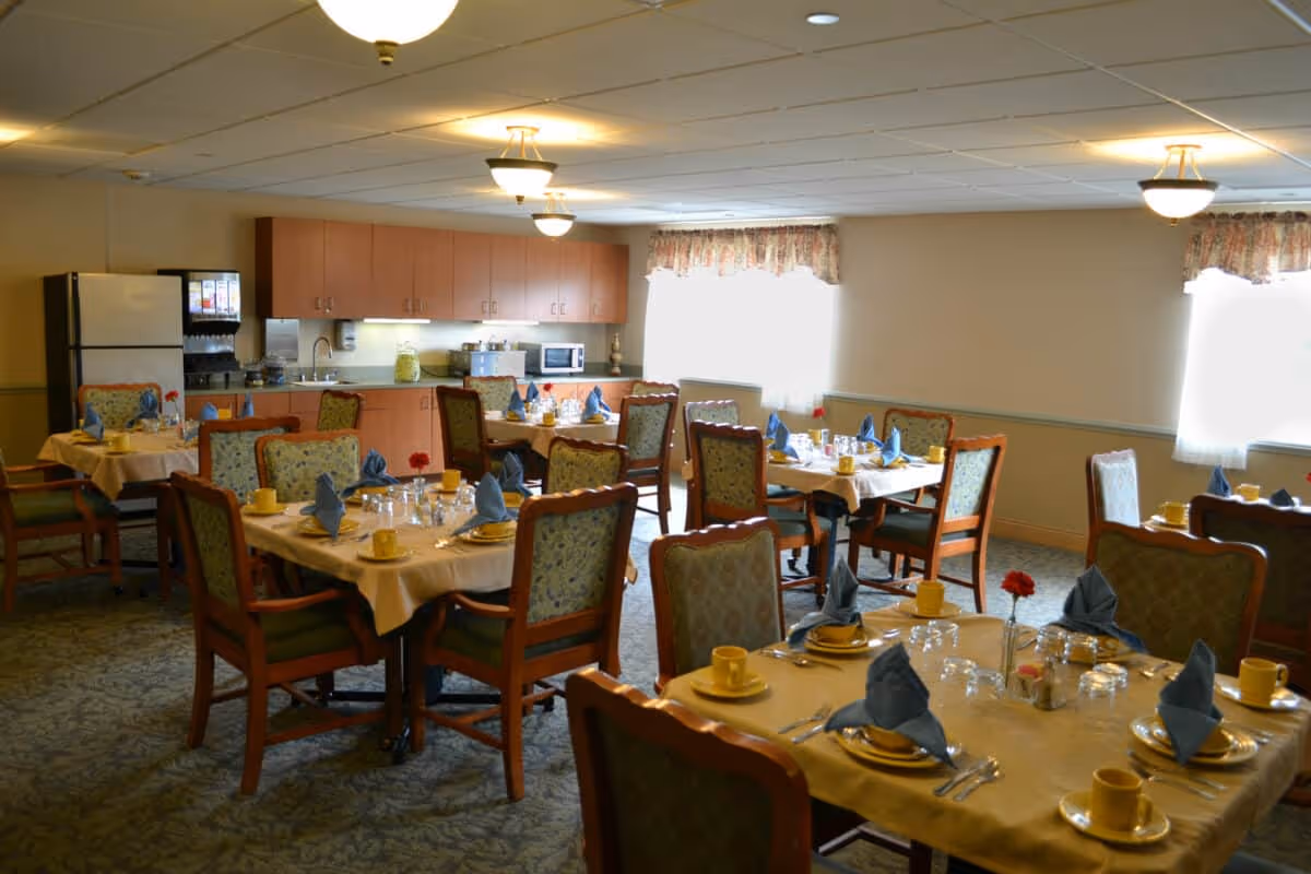 A dining room in a senior living facility with several tables set for a meal. Each table has yellow cups and plates, silverware, glasses, and blue folded napkins. The room has carpeted floors, wooden chairs with cushioned seats and backs, and windows with floral curtains letting in natural light. In the background, there is a kitchenette area with cabinets, a refrigerator, a microwave, and a beverage dispenser.