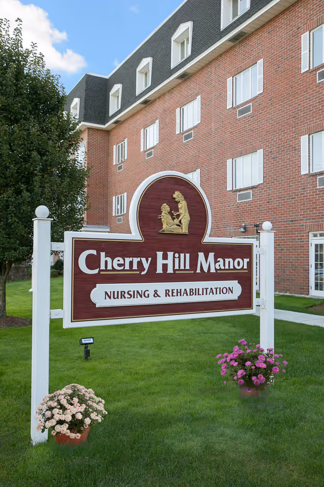 A large maroon and white sign reading 'Cherry Hill Manor Nursing & Rehabilitation' stands on a green lawn in front of a multi-story brick building with white-trimmed windows. There are two flower pots with blooming flowers on either side of the sign and a tree to the left.