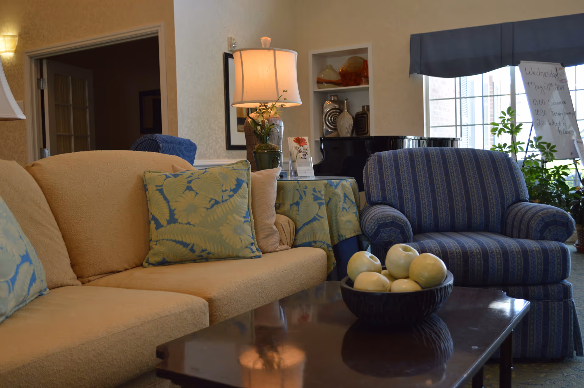 A cozy living room area with a beige sofa adorned with floral patterned cushions, a blue striped armchair, a wooden coffee table with a bowl of decorative white spheres, a table lamp on a side table covered with a floral tablecloth, and a large window with plants and a whiteboard in the background.