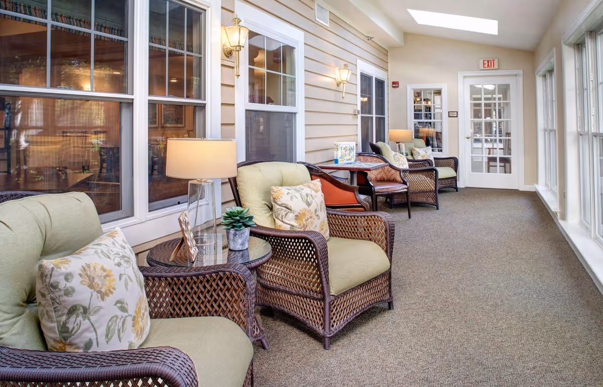 Sunlit enclosed sitting area with wicker armchairs, side tables, lamps, and large windows.