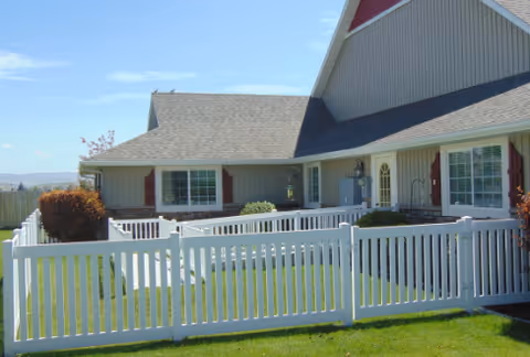 Exterior view of a single-story building with a gray roof and beige siding, featuring multiple windows with red shutters. The building is surrounded by a white picket fence enclosing a green lawn under a clear blue sky.