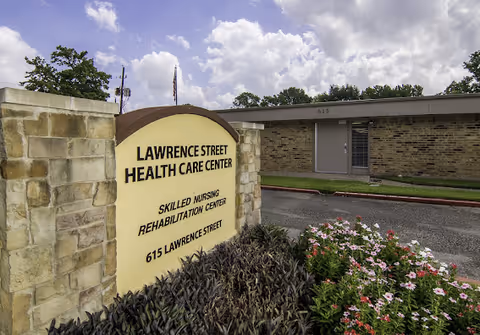 Outdoor view of the entrance area of Lawrence Street Health Care Center, showing a stone sign with the facility name, skilled nursing rehabilitation center, and address 615 Lawrence Street. The building and a cloudy sky are visible in the background, along with some landscaping including flowers and bushes.
