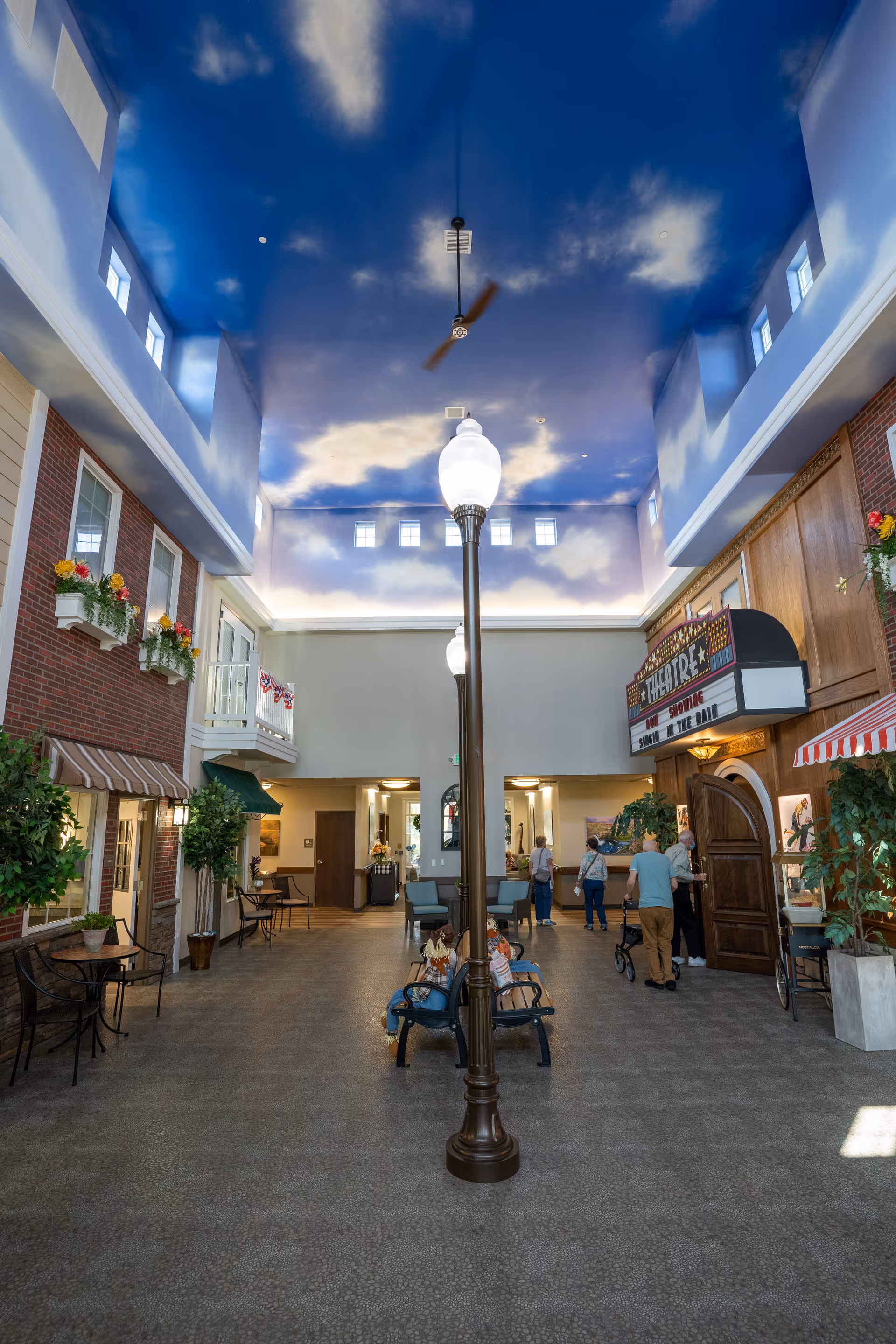Indoor common area of a senior living facility with a high ceiling painted to resemble a blue sky with clouds. The space features a streetlamp-style light fixture in the center, benches, potted plants, and a small theater entrance with a marquee sign. Several people are seen walking and sitting in the area.