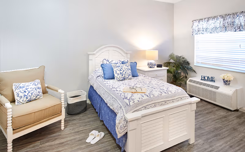 A bright bedroom featuring a white single bed with blue-and-white floral bedding, a beige armchair, nightstand with lamp, window, and slippers on a wood floor.
