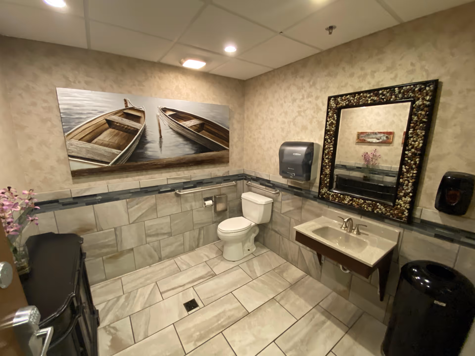 A clean and spacious bathroom with beige patterned walls and light-colored tiled floor and lower walls. The bathroom features a white toilet with grab bars, a wall-mounted sink with a decorative mirror framed with stones, a paper towel dispenser, a soap dispenser, a black trash can, and a small black cabinet with a vase of purple flowers. Above the toilet is a large painting of two wooden boats on water.