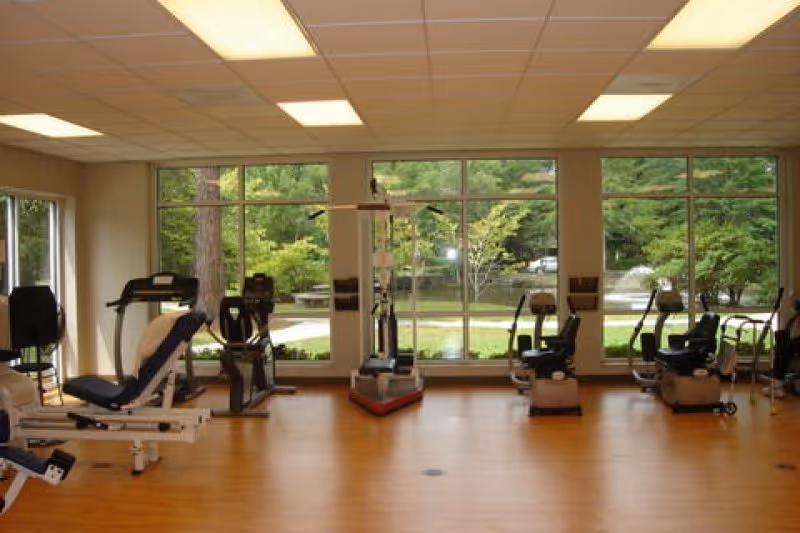 A fitness room with exercise equipment including stationary bikes, a treadmill, and weight machines. Large windows provide a view of a green outdoor area with trees and a fountain.