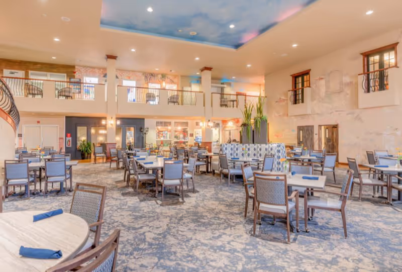 A spacious dining room with multiple tables and chairs arranged neatly. The room features a high ceiling with a sky-themed mural, a second-floor balcony with seating, and large windows letting in natural light. The tables are set with blue napkins, and there are plants and decorative elements throughout the space.