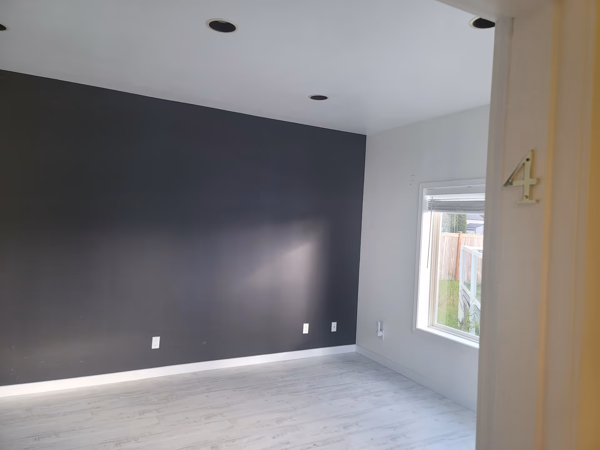Empty room with light-colored wooden floor, a large dark gray accent wall, white ceiling with recessed lights, and a window showing an outdoor view. The door frame on the right side has a number 4 attached to it.