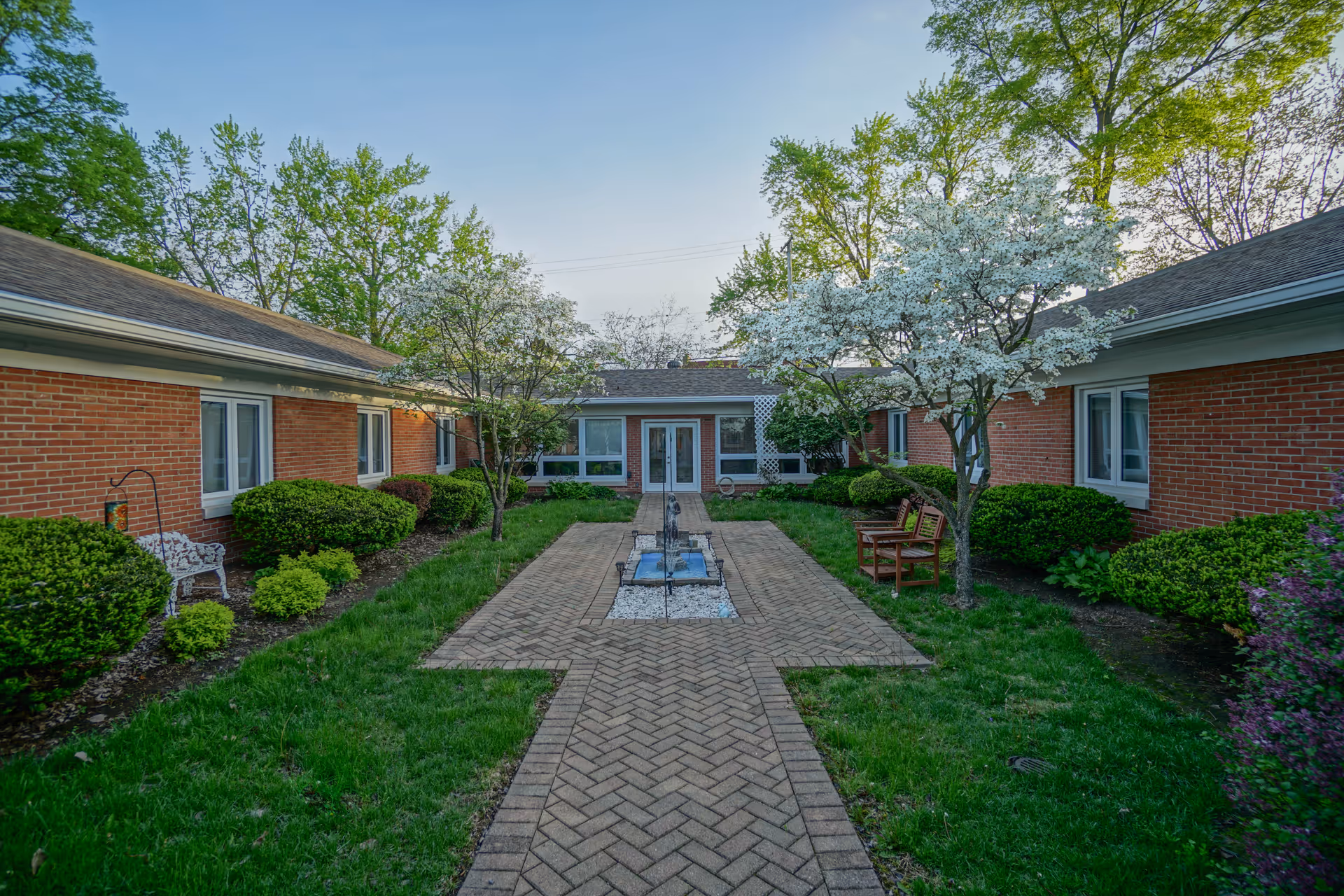 A peaceful outdoor courtyard with a brick pathway leading to a central water feature. The courtyard is surrounded by single-story brick buildings with windows, and there are green bushes and blooming trees with white flowers on either side. Two wooden benches are placed near the trees on the right side.