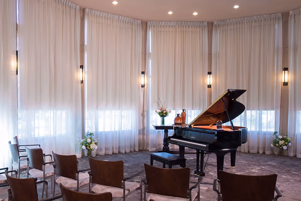 A bright community room with a grand piano at the front, rows of chairs, tall sheer curtains, and floral arrangements.