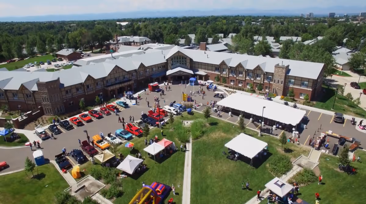 Aerial view of Eastern Star Masonic Retirement Campus showing a large building with a parking lot filled with classic cars and several tents set up on a grassy area, with people walking around and enjoying an outdoor event.