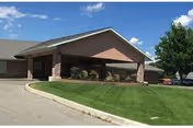 Exterior front entrance of a single-story brick senior living building with a covered porte-cochère, driveway, and manicured lawn under a blue sky.