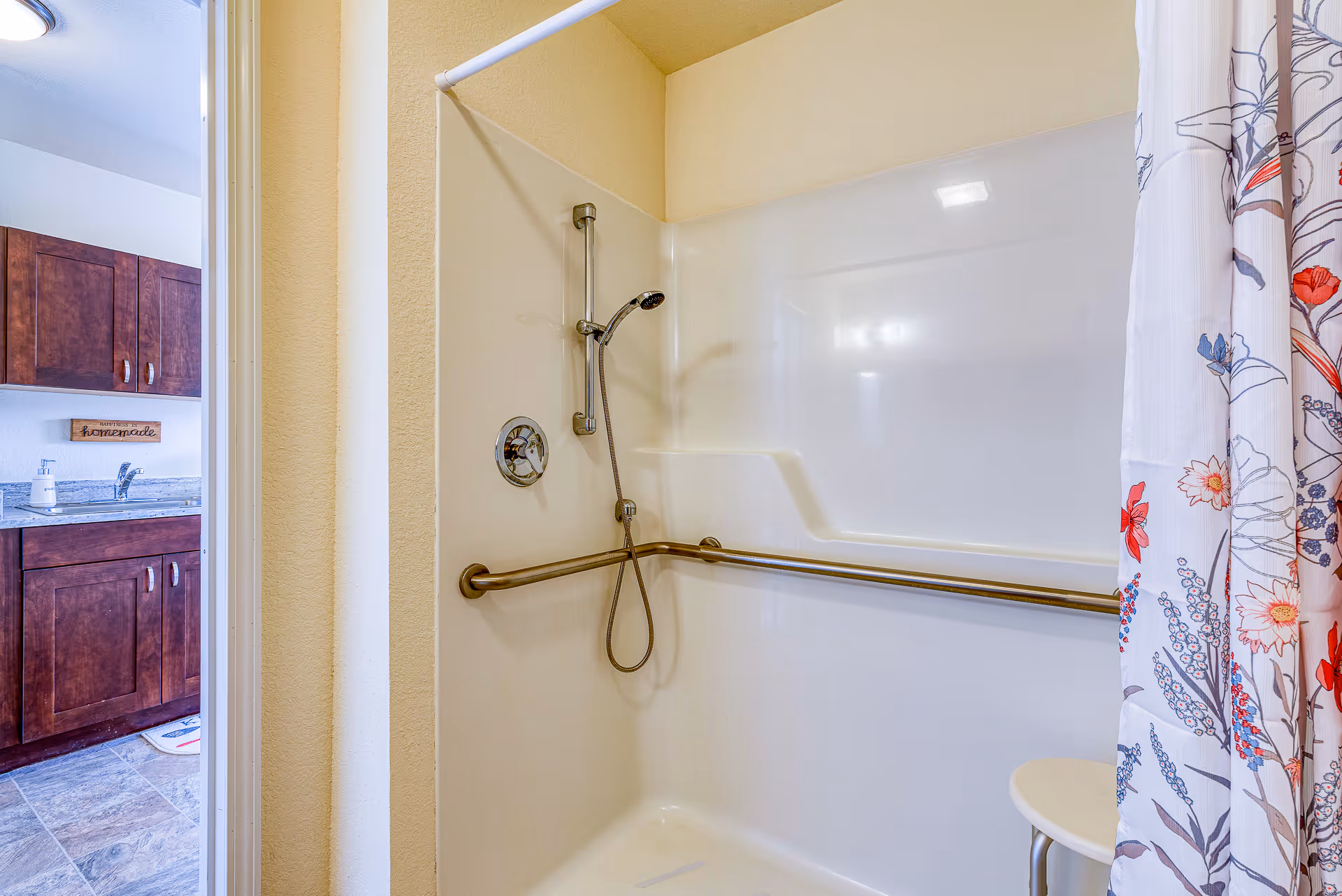Shower stall with a horizontal grab bar, handheld showerhead and floral curtain, with a sink and wooden cabinets visible through the open doorway.