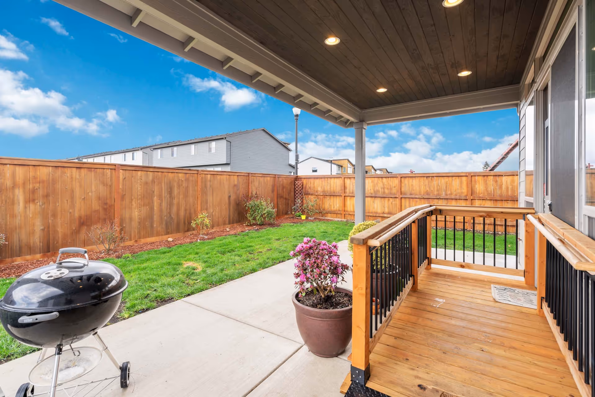 Covered wooden patio with railing overlooking a fenced backyard with green grass, small plants, and a charcoal grill on a concrete patio. The sky is blue with some clouds, and neighboring houses are visible beyond the wooden fence.