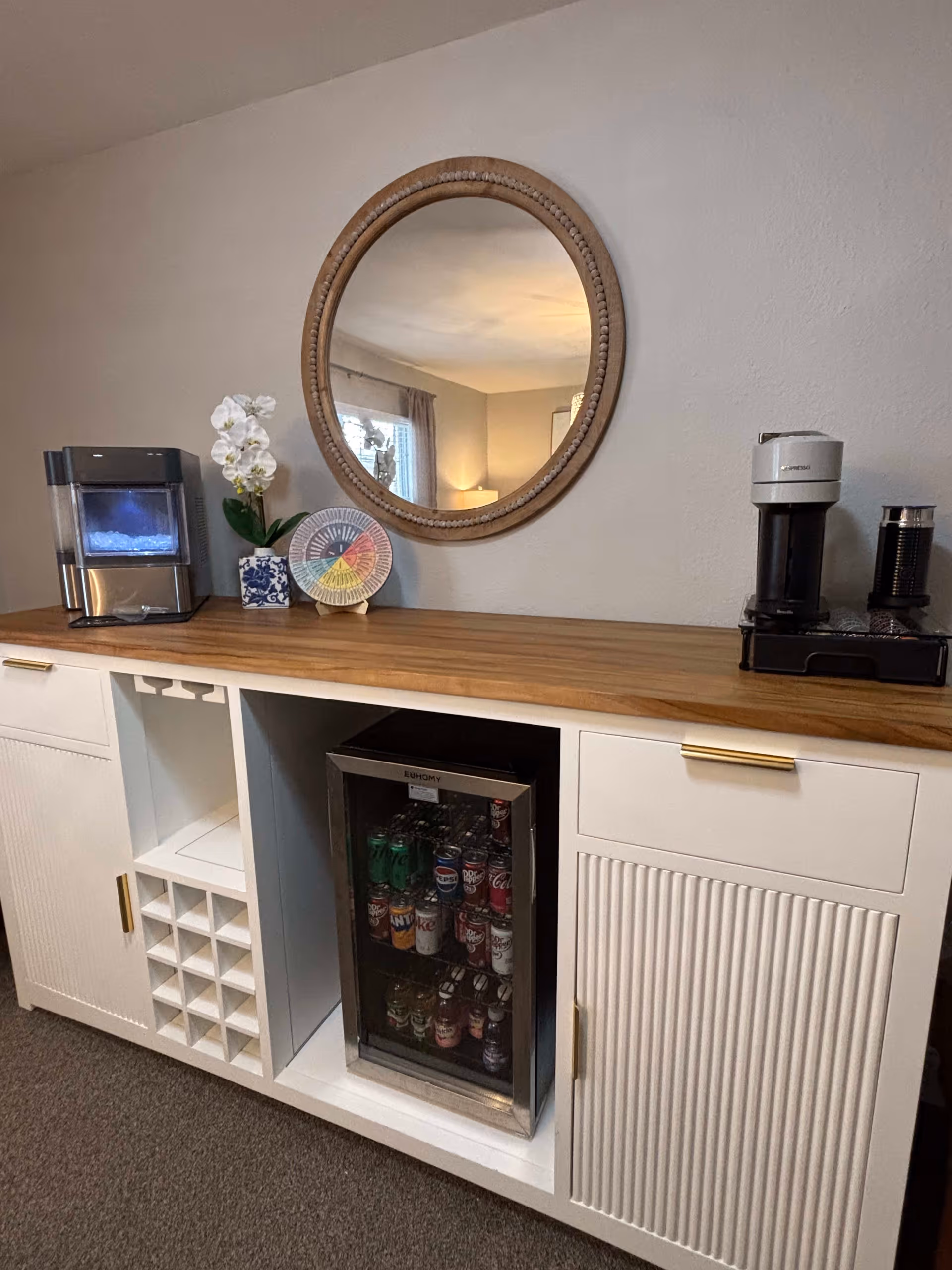A wooden countertop with a white cabinet below it. The cabinet has a built-in mini fridge stocked with various canned drinks, a wine rack, and closed storage compartments. On the countertop, there is a small ice maker, a white orchid plant in a blue and white pot, a colorful circular decorative item, and a coffee maker. A round mirror with a wooden frame hangs on the wall above the countertop, reflecting part of the room with a window and a lamp.