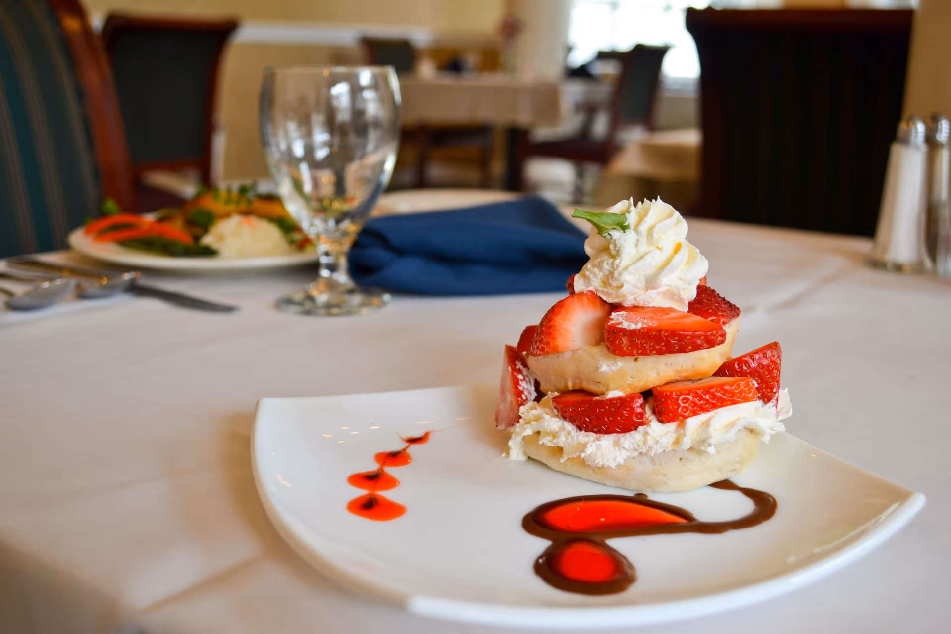 A plated strawberry shortcake dessert with sauce on a white plate set on a linen-covered dining table with glassware and chairs in the background.