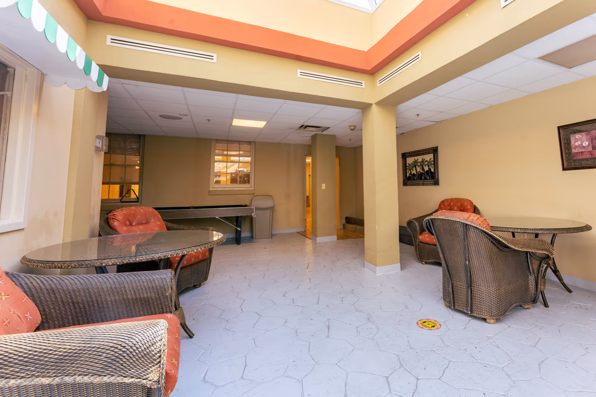 Indoor seating area with wicker chairs and glass-top tables, featuring red cushions. The room has beige walls, a tiled floor, and a skylight allowing natural light. There are framed pictures on the walls and a doorway leading to another room.