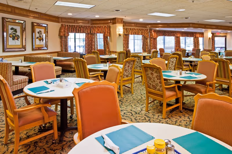 A spacious dining room with multiple round tables set with teal placemats, napkins, and silverware. The room features upholstered chairs in warm tones, patterned carpet, and large windows with floral curtains allowing natural light to fill the space. Framed botanical artwork decorates the walls.
