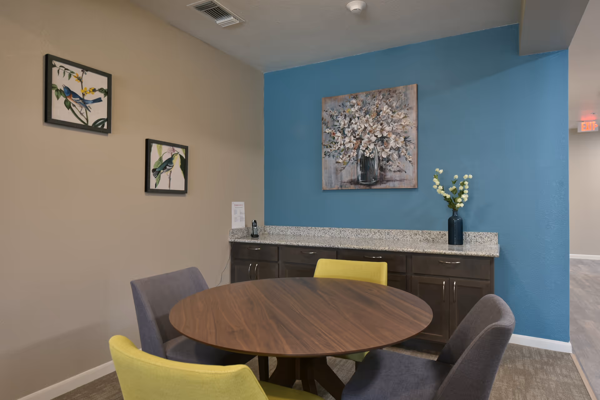 A small dining area with a round wooden table surrounded by four chairs, two yellow and two gray. Behind the table is a granite countertop with dark cabinets underneath. On the wall above the countertop is a large painting of white flowers in a vase, and on the adjacent wall are two smaller framed pictures of birds. The walls are painted beige and blue.