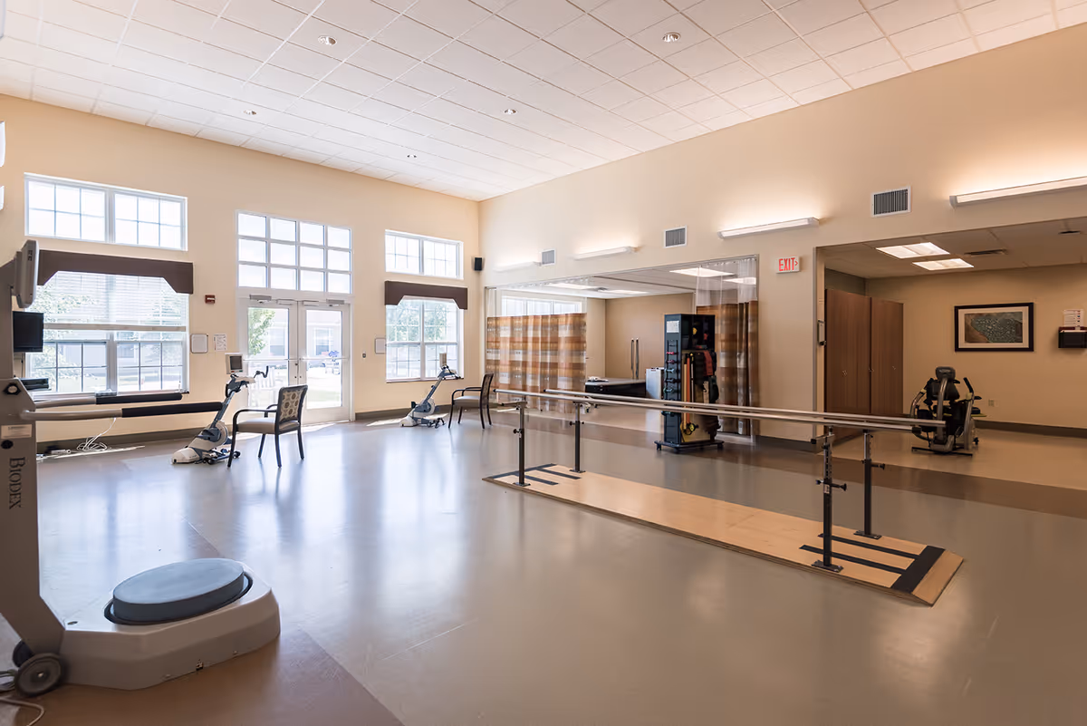 Bright rehabilitation/therapy room with parallel walking bars, exercise equipment, chairs, and large windows.