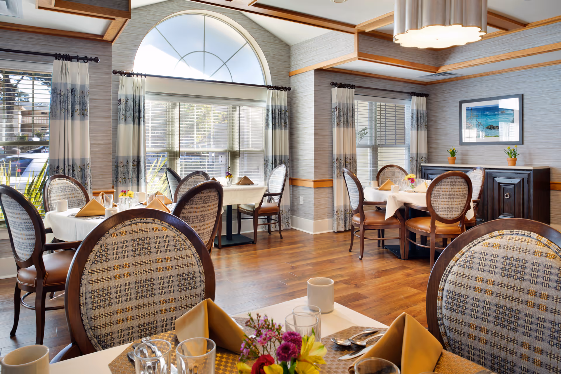 A bright and elegant dining room with round tables covered in white tablecloths, set with beige napkins, glasses, and silverware. The room features large windows with patterned curtains, wooden floors, and a decorative ceiling light fixture. There are framed pictures and small potted plants on a sideboard against the wall.