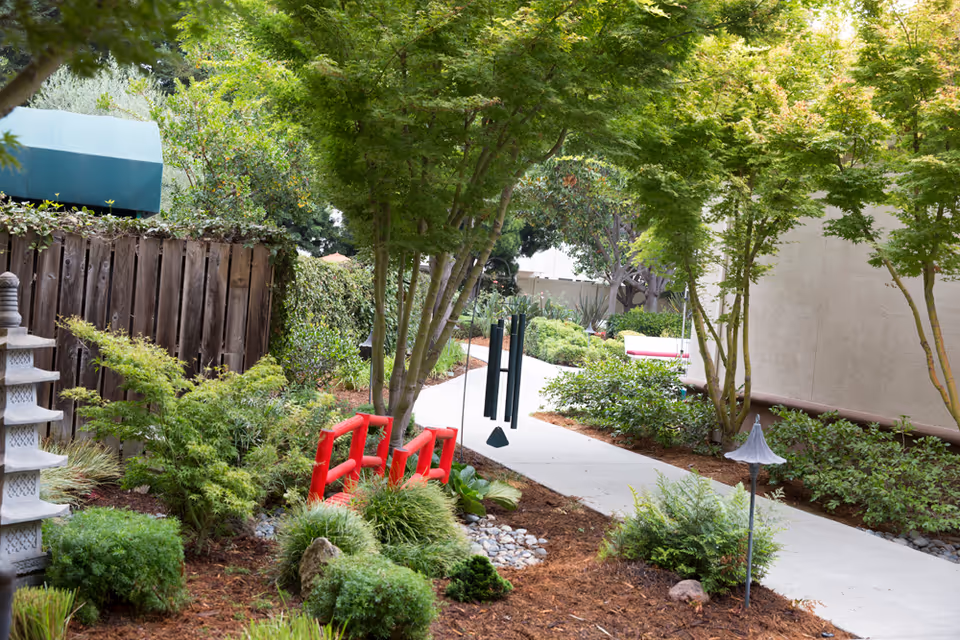Courtyard garden with a paved walkway, trees and shrubs, a small red decorative bridge and hanging wind chimes.