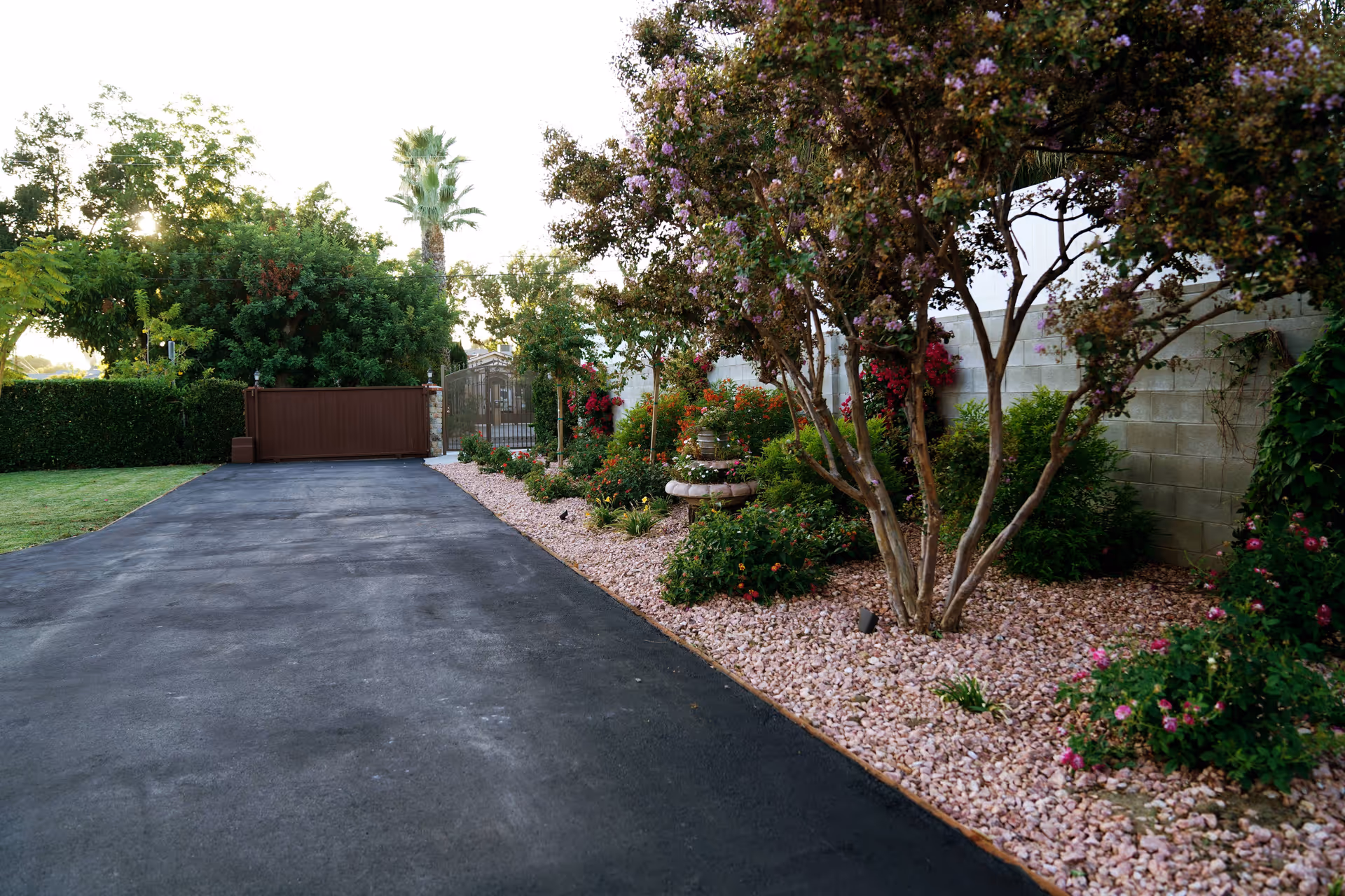 A paved driveway leading to a closed brown gate with a garden bed on the right side filled with flowering bushes, small trees, and a stone birdbath. There is a concrete block wall behind the garden and various green trees and plants in the background.