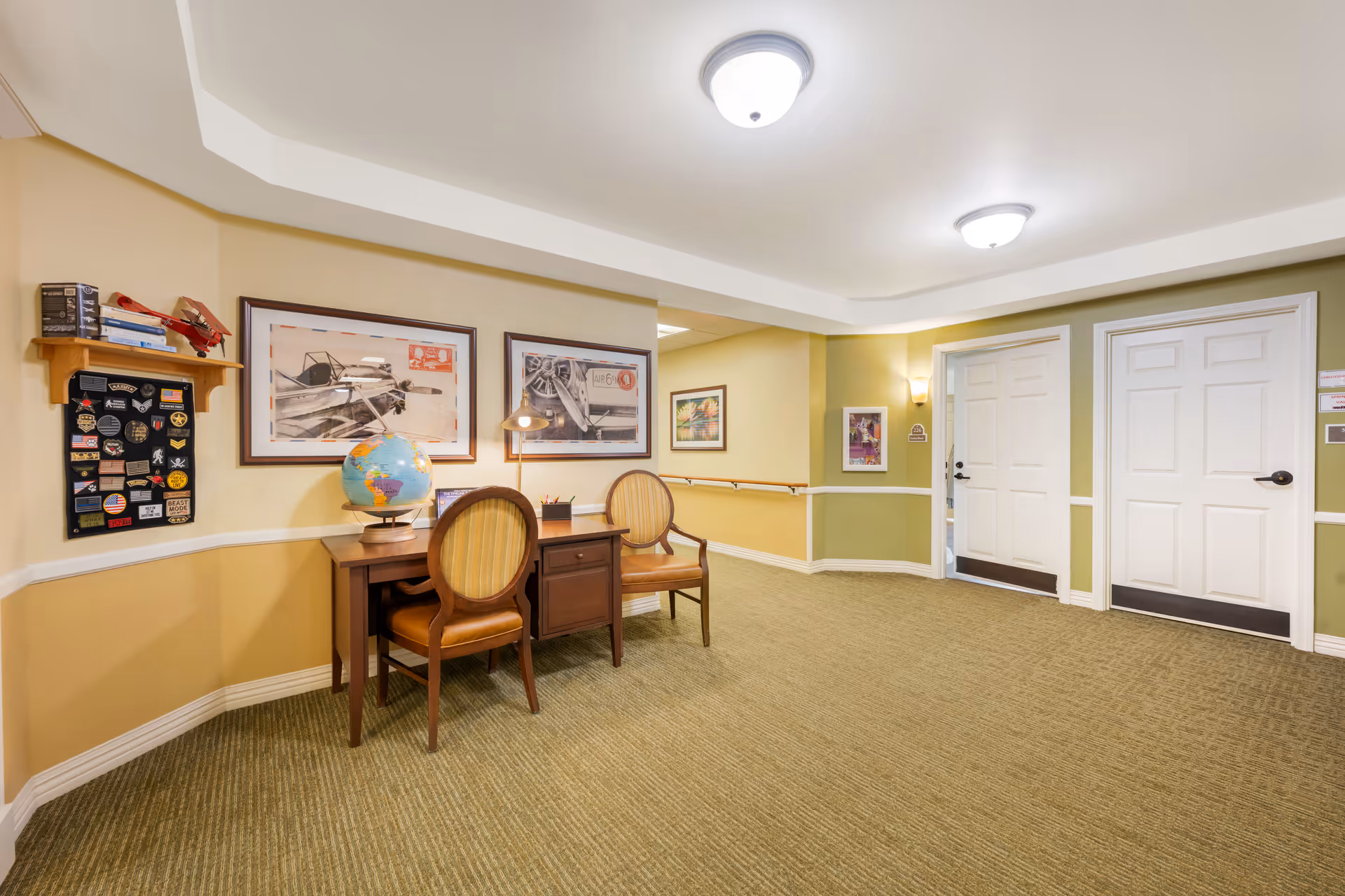 Well-lit senior living interior with a small desk and two chairs, a globe and framed artwork along a carpeted corridor with two white doors.