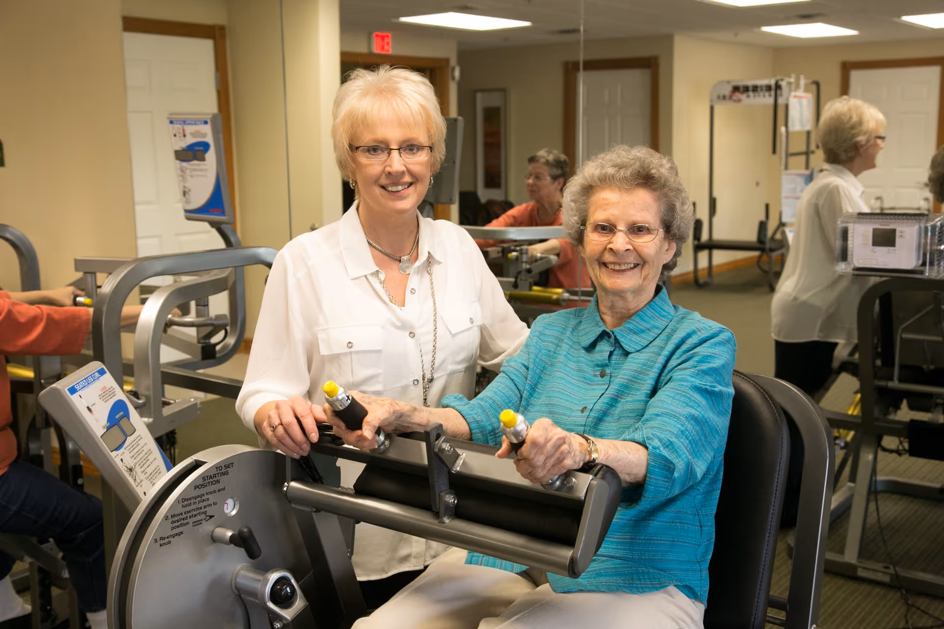 Two elderly women smiling in a fitness room at a senior living facility. One woman is seated on an exercise machine holding the handles, while the other woman stands beside her, offering support. The room has exercise equipment and mirrors on the walls.