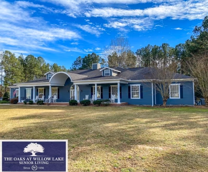 Single-story blue senior living building with a covered front porch and rocking chairs on a large lawn under a blue sky.