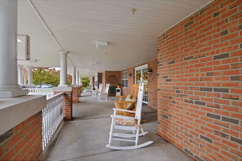 Covered outdoor porch area with white rocking chairs and a wooden bench along a brick wall, overlooking a landscaped area with trees.