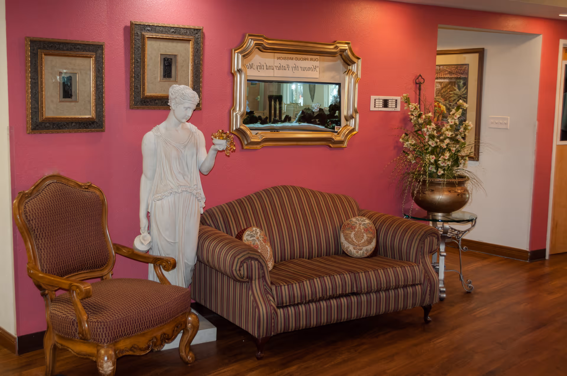 A cozy seating area with a striped loveseat and a wooden armchair with patterned upholstery. A white classical statue holding a small bowl stands between the chairs. The wall behind is painted pink and decorated with two framed pictures and a large ornate gold-framed mirror. A large bronze vase with flowers sits on a glass-topped side table next to the loveseat. The floor is wooden.
