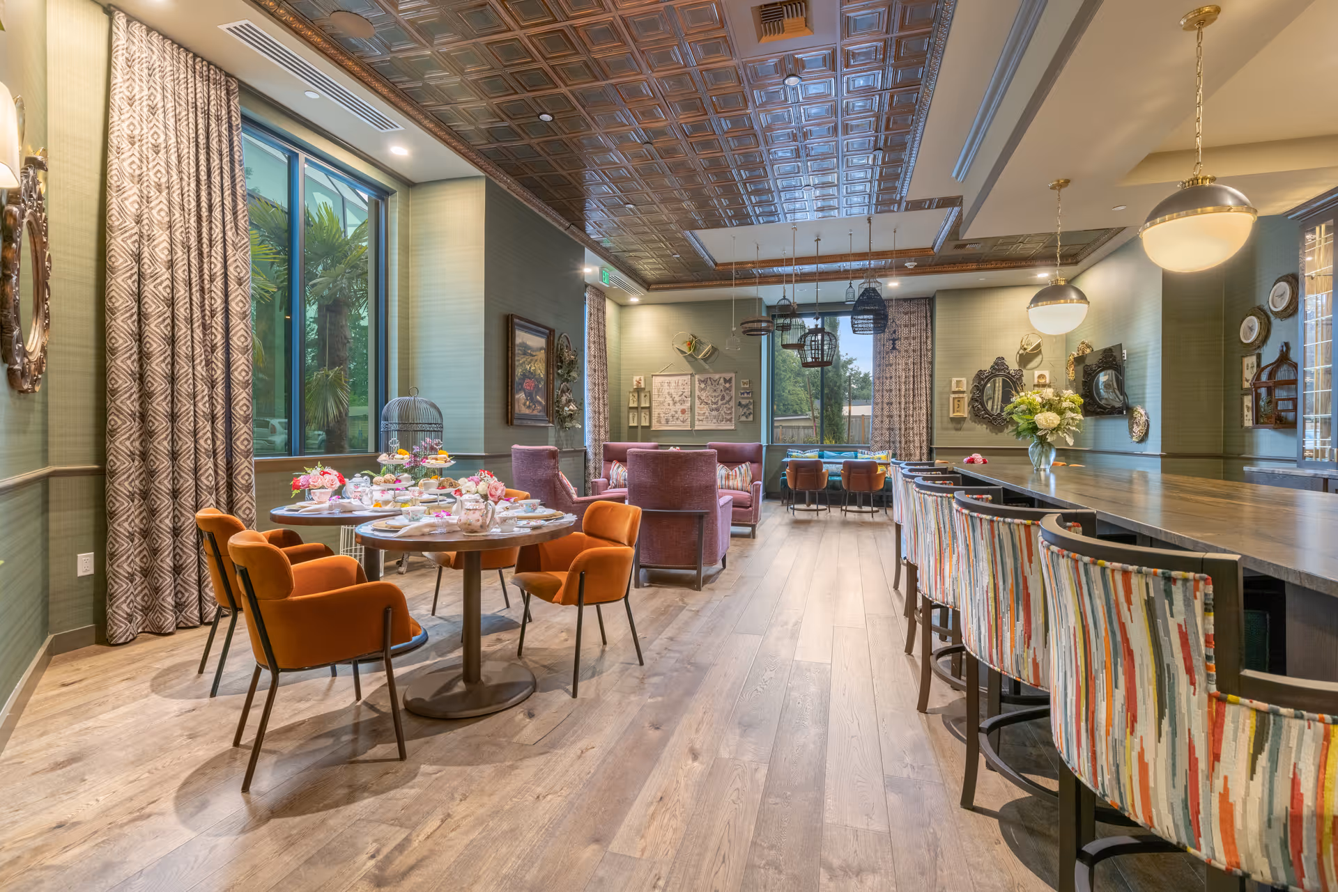 Elegant dining and lounge room with tables set for tea, orange upholstered chairs, a long bar with patterned stools, and large windows.