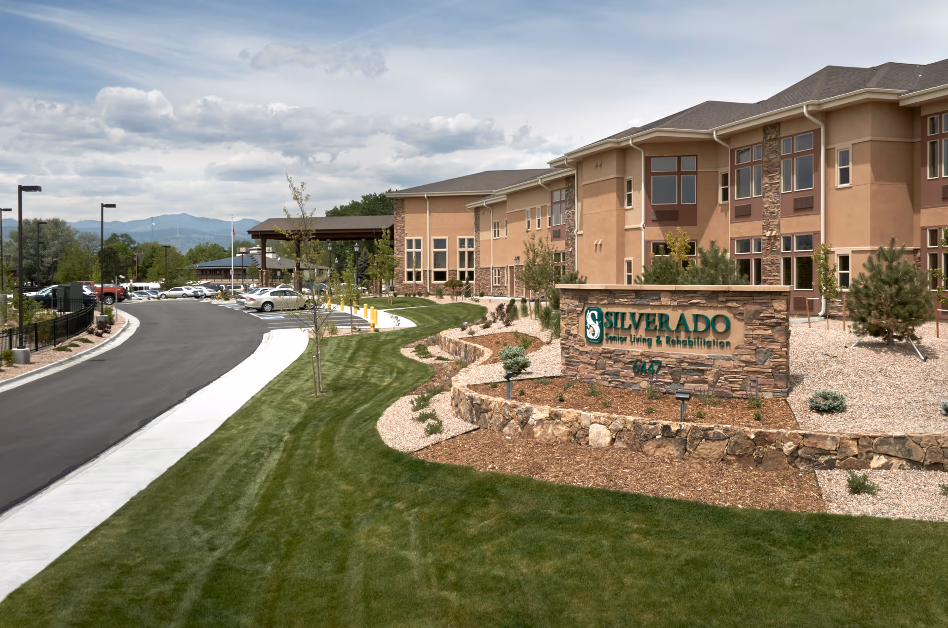 Exterior view of a senior living and rehabilitation facility named Silverado, showing a large building with multiple windows, a stone sign with the facility name, a well-maintained lawn, landscaped garden beds, a driveway, and parked cars under a cloudy sky.