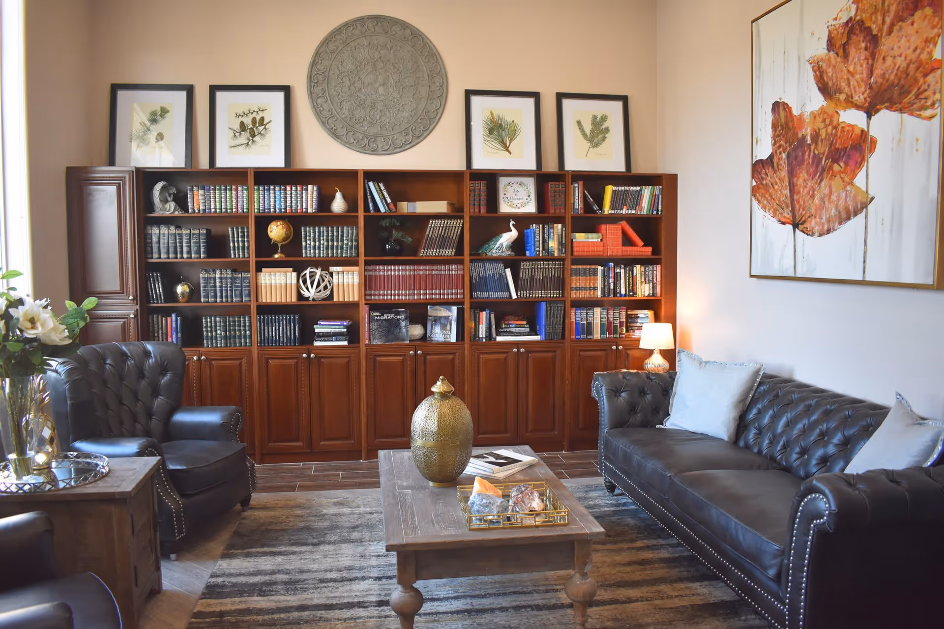 A cozy living room with a dark leather sofa and matching armchairs surrounding a wooden coffee table. Behind the seating area is a large wooden bookshelf filled with books and decorative items. The walls are adorned with framed botanical prints and a large painting of orange flowers. A small table lamp provides warm lighting.