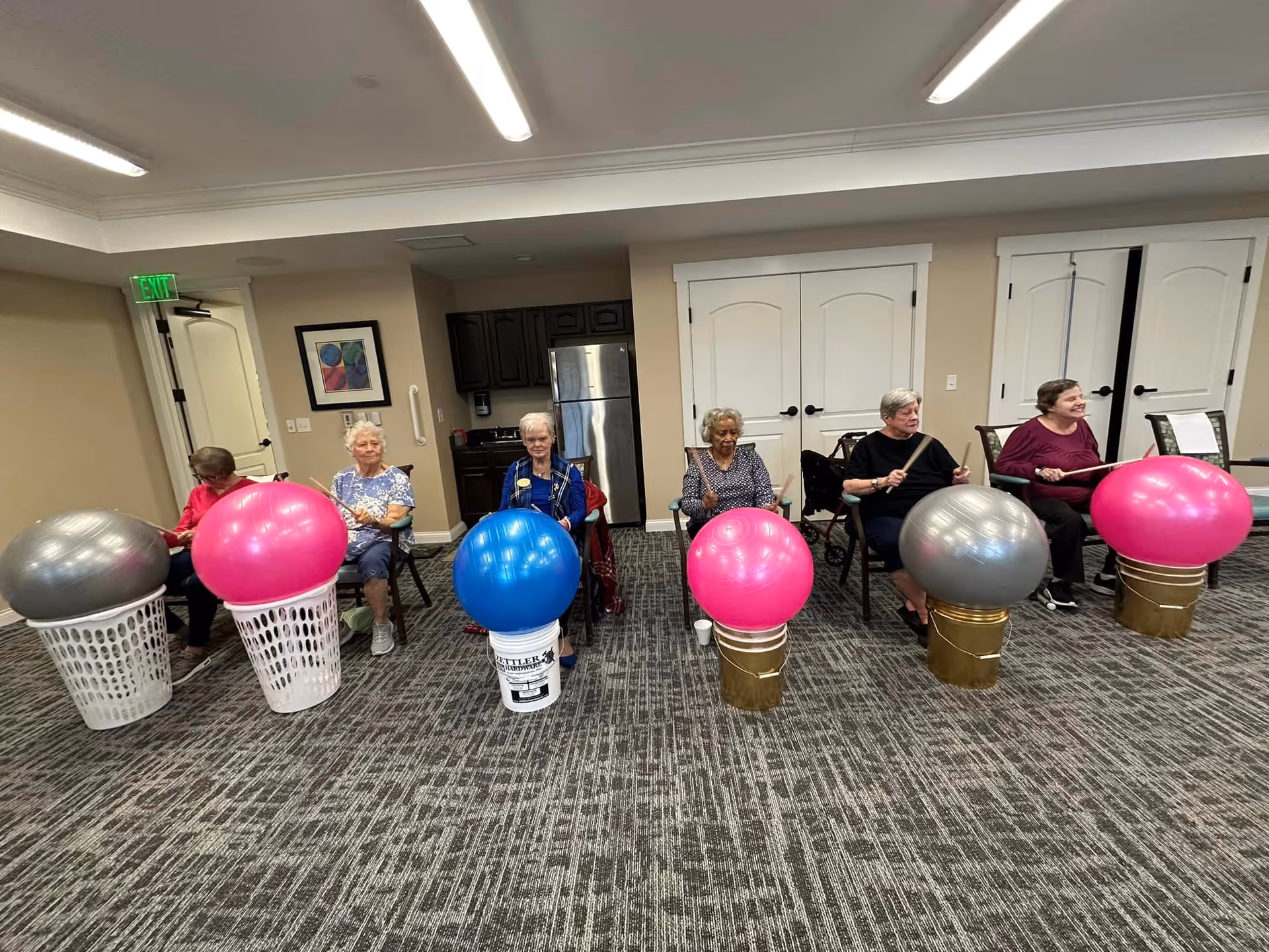 A group of six elderly women seated in a room, each with a large exercise ball placed on a laundry basket or bucket in front of them. They appear to be participating in a group exercise activity using drumsticks to hit the balls. The room has carpeted flooring, beige walls, a refrigerator, and white double doors in the background.