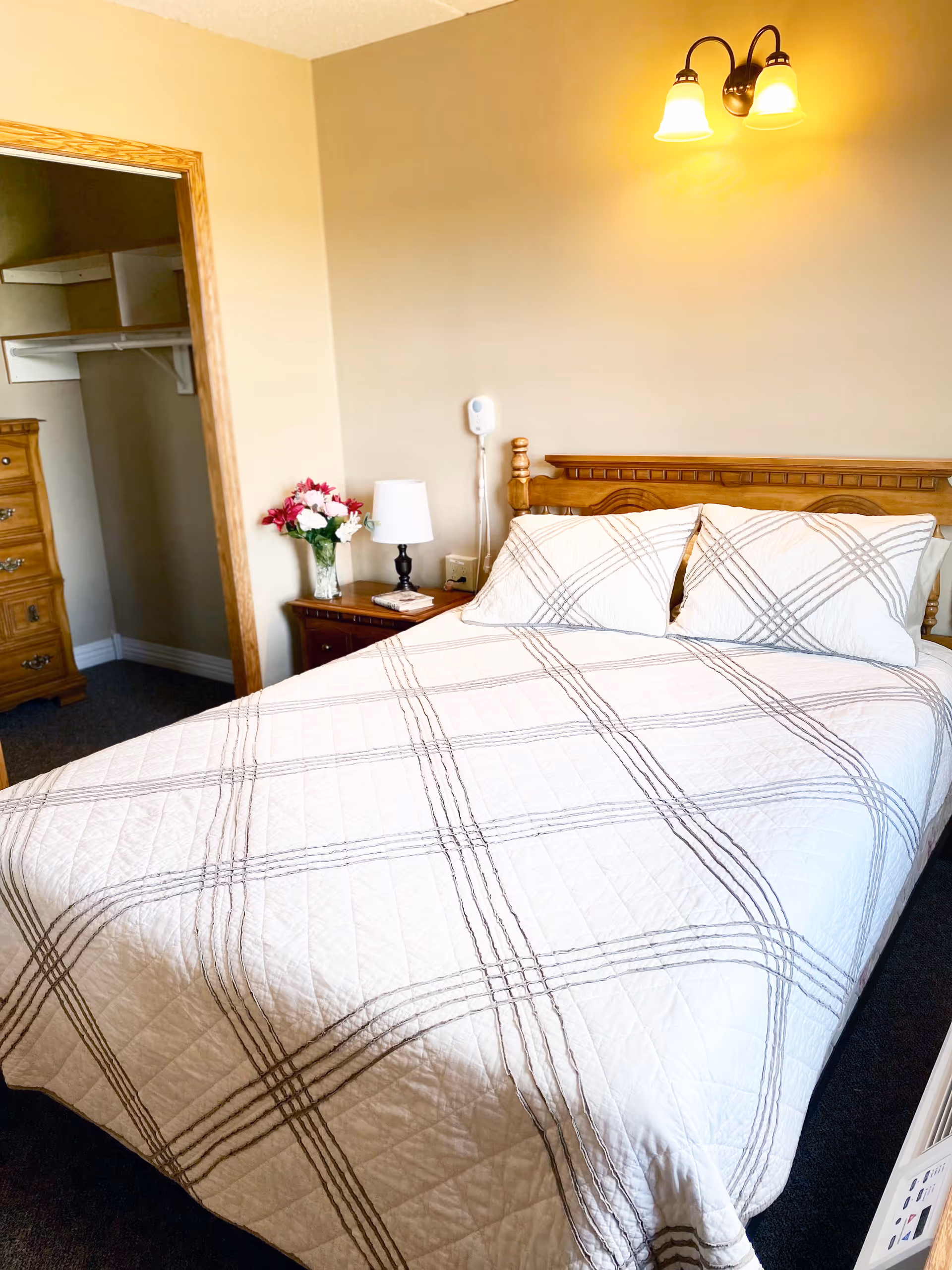 A bedroom with a neatly made bed featuring a white quilt with a gray plaid pattern. There is a wooden headboard, a nightstand with a white lamp and a vase of flowers, and an open closet with wooden drawers visible. A wall-mounted light fixture with two lamps is above the bed.