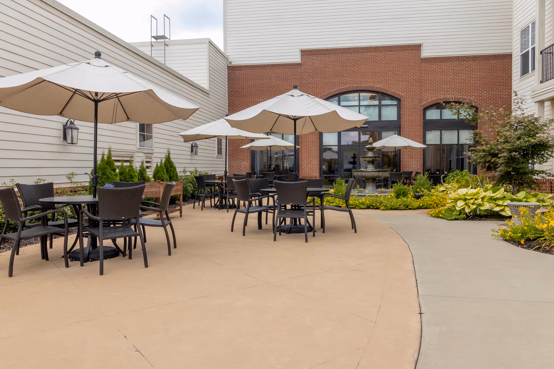 Outdoor patio area with several tables and chairs under large beige umbrellas. The patio is surrounded by plants and flowers, with a multi-tiered water fountain in the background near a brick building with large windows.