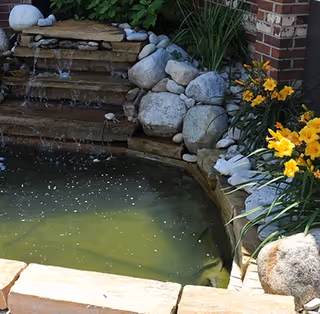 A small outdoor pond with a cascading waterfall surrounded by large rocks and yellow flowers next to a brick wall.